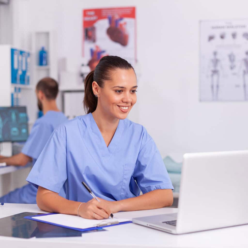 Medical practitioner sitting at desk in hospital office using laptop with doctor in the background. Health care physician using computer in modern clinic looking at monitor, medicine, profession.